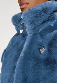 Casaco de pelo sintético em azul profundo, com colarinho alto, fecho de correr com puxador e um logo da EA no lado esquerdo.