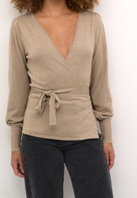 Beige wrap top with a deep V-neck, long puff sleeves, and a tied waist detail. Soft knit fabric, smooth texture, paired with dark jeans.