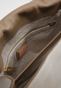Intérieur d'un sac à main en cuir tan avec une doublure beige, une fermeture éclair dorée et une étiquette de marque. Matériau doux et texturé avec un design décontracté.