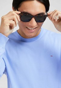 Pull en tricot bleu clair avec un col rond, arborant un petit logo rose sur la poitrine. La personne porte des lunettes de soleil sombres.