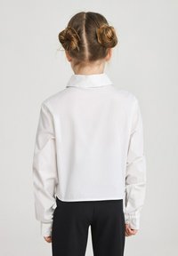 Blusa bianca a maniche lunghe con colletto e polsini abbottonati, caratterizzata da un orlo corto e una texture liscia. Indossata con pantaloni neri.
