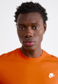 Oranžs kokvilnas T-krekls ar apaļu kaklu un īsām piedurknēm. Uz kreisā krūšu kurvja ir neliels balts Nike logo. Gluda tekstūra.