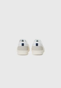 Zapatillas blancas con acentos en el talón de color gris y una etiqueta azul marino en la parte trasera, exhibidas sobre un fondo blanco liso.