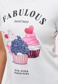 Λευκό πουκάμισο με πολύχρωμη εικονογράφηση με κεκάκια, διακόσμηση με παγέτες και κείμενο που λέει «Υπέροχη γλύκα, δαγκωνιά τελειότητας σε μπουκίτσες.»