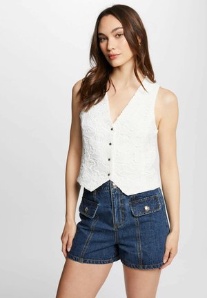 Donna con gilet bianco senza maniche in pizzo abbottonato e pantaloncini di jeans blu a vita alta con tasche a pattina frontali, in piedi davanti a uno sfondo neutro.