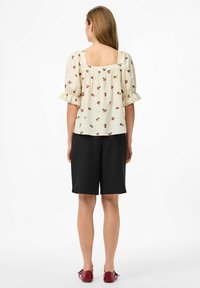 Blusa floreale a maniche corte color avorio con motivo a ciliegie rosse, scollo quadrato e maniche arricciate, abbinata a pantaloni corti neri e scarpe rosse.