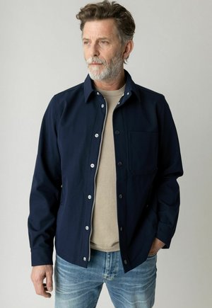 Man with gray beard wearing unbuttoned navy jacket over beige shirt and blue jeans, standing against plain light background, looking left.