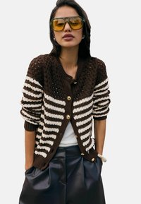 Brązowo-kremowy kardigan w paski z fakturowanym splotem, ozdobiony złotymi guzikami i ściągaczami na rękawach. Noszony na białej bluzce i czarnych spodniach.