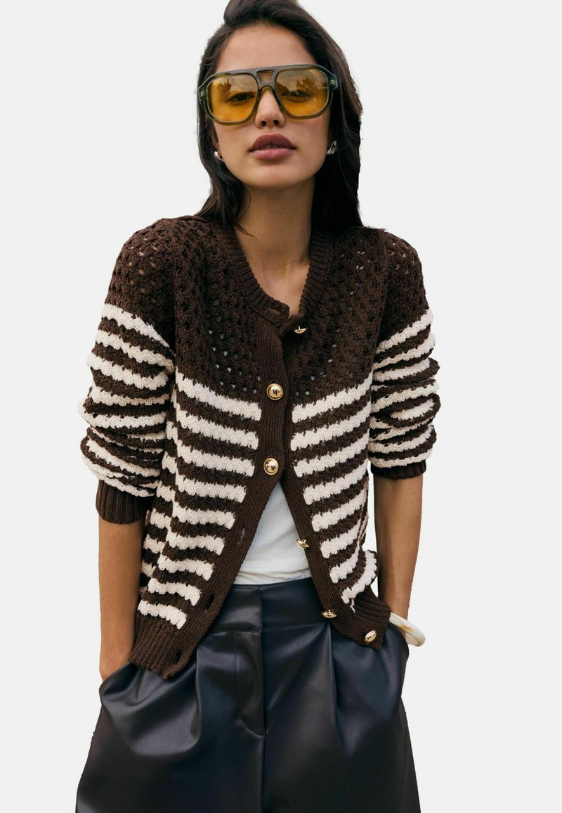 Brązowo-kremowy kardigan w paski z fakturowanym splotem, ozdobiony złotymi guzikami i ściągaczami na rękawach. Noszony na białej bluzce i czarnych spodniach.