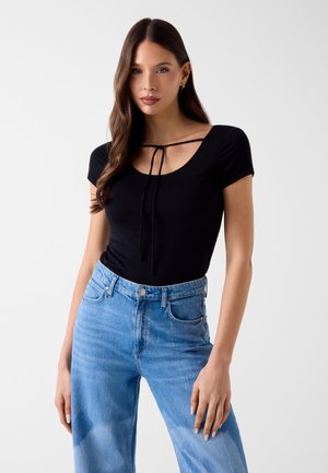 Kobieta o długich brązowych włosach, ubrana w czarny krótki top z wiązaniem z przodu i jasnoniebieskie dżinsy z wysokim stanem, stojąca.