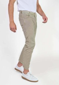 Khaki cargobroek met een tapered fit, gemaakt van duurzaam materiaal, heeft zijzakken en een ritsluiting, gedragen met witte sportschoenen.
