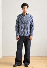 Ναυτικό πουκάμισο με λουλούδια και λευκό περίγραμμα, συνδυασμένο με τζιν παντελόνι με φαρδιά πόδια σε σκούρο χρώμα και μαύρα παπούτσια, στέκεται σε ξύλινο πάτωμα.