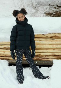 Schwarze Pufferjacke, gemusterte schwarze Hose mit weißen Blumen und passende Handschuhe. Steht auf Schnee mit Holzstämmen im Hintergrund.
