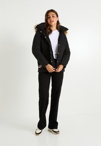 Chaqueta acolchada negra con capucha, con ribete de piel sintética, cierre de cremallera y detalles en tono dorado. Combinada con pantalones negros y zapatillas multicolor.