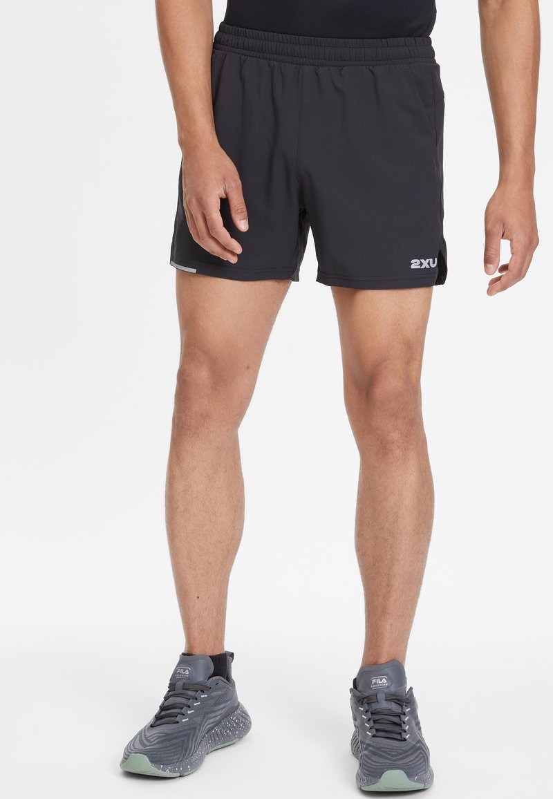 Pantalones cortos deportivos negros con cintura elástica, aberturas laterales y un pequeño logotipo en el muslo. Combinados con zapatillas gris texturizadas.