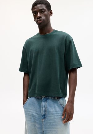 T-shirt en coton vert foncé avec des manches courtes, col rond et coupe décontractée. Porté avec un pantalon en denim bleu clair, à taille haute et coupe ample.