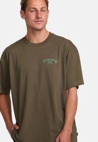 Olive green t-shirt made of cotton with a crew neck and short sleeves. Features a subtle logo design on the chest. Simple, relaxed fit.