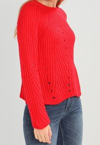 Un pull en tricot rouge avec une texture côtelée, un col rond et de petites découpes. Associé à un jean en denim bleu.