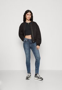 Chaqueta bomber negra, top corto negro, jeans ajustados azules y zapatillas de running negras con acentos blancos. Fondo plano y de larga extensión.