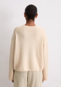 Pull oversized beige clair en laine texturée, avec des poignets et un col côtelés. Porté avec un pantalon en velours côtelé de couleur claire.