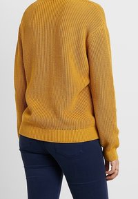 Musztardowo-żółty sweter z dzianiny o prążkowanej fakturze, długich rękawach i luźnym kroju. Posiada szeroki okrągły dekolt i lekko skrócony dół.