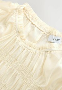 Svijetlo kremasta bluza s nabranim strukom i naboranim izrezom. Ima teksturu nabrane tkanine i etiketu s natpisom "NEXT".