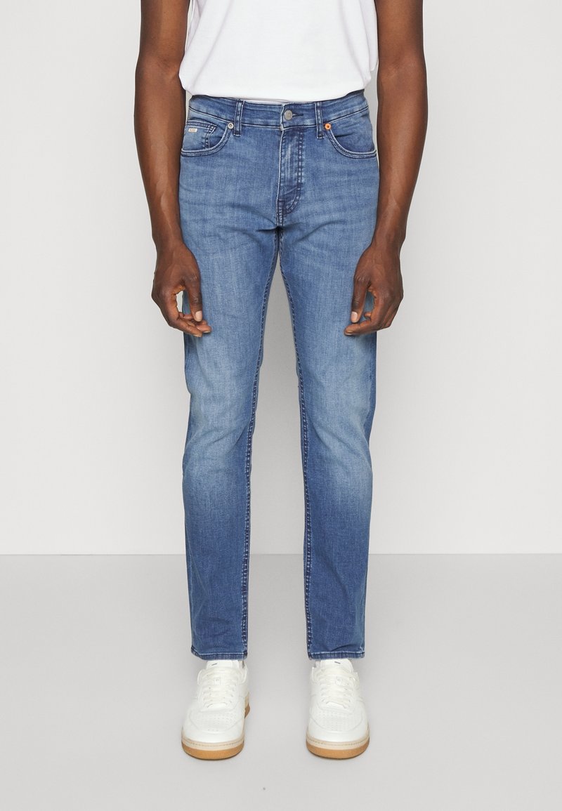 Man in mediumblauwe slim-fit jeans en witte sneakers die tegen een effen lichte achtergrond staat.