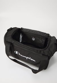 Svart gymbag med glidelås på toppen, justerbar stropp og hvit "Champion" logo på siden. Innvendig med lomme og mykt fôr.