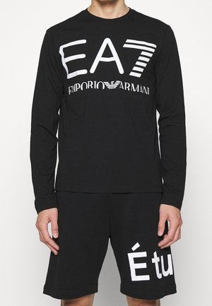 Mann trägt ein schwarzes Langarmshirt mit dem "EA7 Emporio Armani"-Logo und schwarze Shorts mit großem, teilweise weißem Text auf dem rechten Bein.