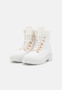 Par af hvide læder vinterstøvler med kraftige såler, gyldne øjer, hvide snørebånd og for med fleece på en ensfarvet baggrund.