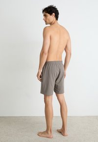 Grå bomullsshorts med elastisk midje, knelengde, synlige sømmer og en glatt tekstur. Modell vender bort fra kameraet.