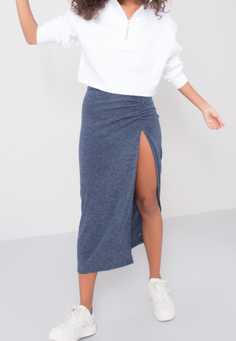 Jupe bleu marine, longueur genoux avec fronces sur les côtés et une fente haute, associée à un sweat-shirt cropped blanc et des baskets blanches.