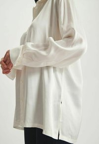 Chemisier blanc à manches longues avec un devant à boutons, texture lisse, coupe décontractée et fentes sur les côtés, fabriqué en tissu léger.