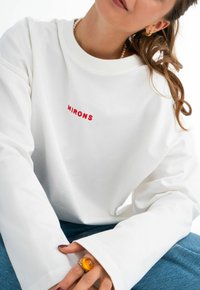 Weißes Sweatshirt mit rotem gesticktem Text "MIRONS", mit weit geschnittenen Ärmeln und einer weichen Textur. Begleitet von blauen Jeans und einem gelben Ring.