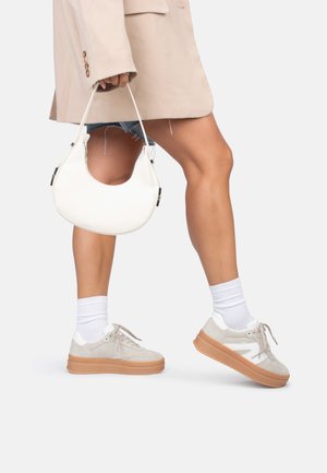 Zapatillas de ante gris claro con detalles blancos y una suela de plataforma en tono beige. Un bolso blanco de forma curvada cuelga de una mano que lleva una blazer beige de gran tamaño.