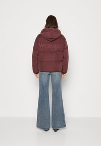 Bordowa kurtka puchowa z kapturem, z logo "Wrangler" na plecach, zestawiona z dzwonowymi niebieskimi dżinsami i czarnymi botkami za kostkę.