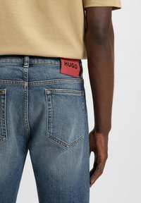 Jeans in denim di colore azzurro chiaro con una texture sbiadita. Presentano un patch in pelle rossa etichettato "HUGO" sulla vita e due tasche posteriori.