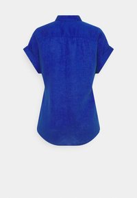 Blå kortärmad skjorta med uppvikta ärmar, krage och rundad nederkant, visad bakifrån mot en enkel vit bakgrund.