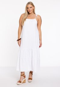 Weißes Baumwoll-Maxi-Kleid mit einem gestuften Saum, verstellbaren Spaghettiträgern und einer gerafften Taille. Ein Paar braune Keilsandaletten vervollständigt den Look.