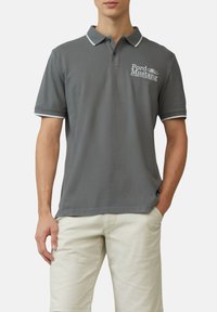 Grijze polo shirt van katoen, met een kraag met blauwe bies en een witte Ford Mustang-logo op de linkerborst. Korte mouwen.
