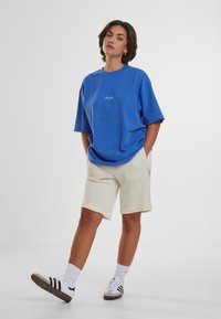 Blå oversized t-skjorte med brodert tekst, parret med kremfargede teksturerte shorts og hvite joggesko med svarte striper og hvite sokker.