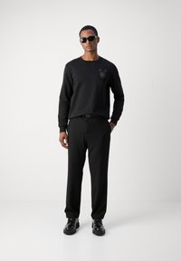Sudadera negra con diseño texturizado en el pecho, combinada con pantalones negros. Modelo que lleva gafas de sol negras y zapatos negros, de pie sobre un fondo liso.