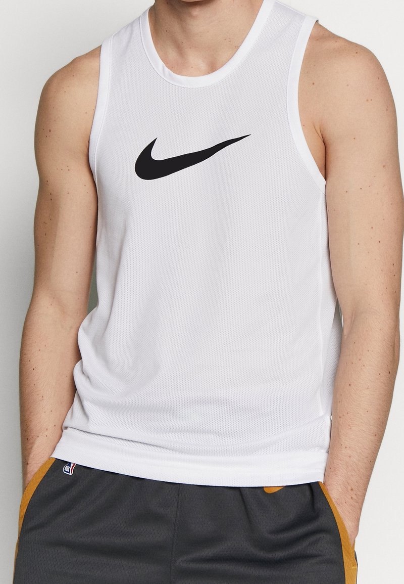 Homme portant un débardeur de sport blanc sans manches Nike et un short gris foncé avec une bande orange, debout les mains dans les poches.