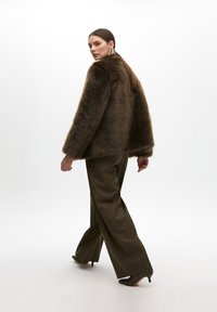 Brown fur coat with a loose fit, paired with olive green wide-leg trousers and black heeled shoes. The model is shown in a turned posture.