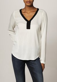 Blouse blanche avec une finition texturée, dotée d'un col en V noir et d'une patte de boutonnage. Manches longues et ourlet arrondi complètent le design.