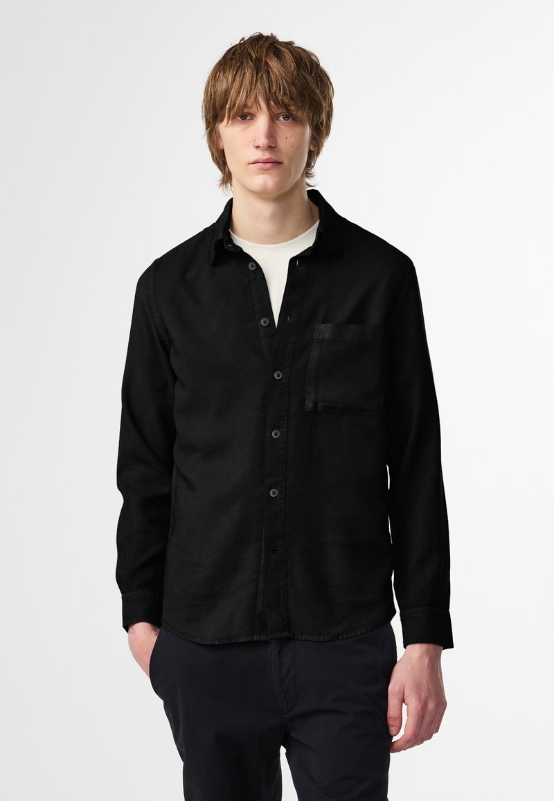 Camicia nera con bottoni, maniche lunghe e una tasca sul petto, realizzata in un tessuto liscio. Indossata sopra una maglietta bianca a girocollo.