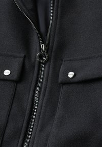 Giacca nera di lana con zip centrale in argento e due tasche a patta chiuse da bottoni metallici rotondi.