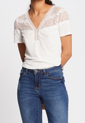 Vrouw draagt een wit shirt met korte mouwen, kant inzetstuk en V-hals, gecombineerd met blauwe spijkerbroek, staand met handen achter de rug.