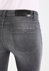 Jeans de mezclilla gris con un ajuste ceñido, que presentan un bolsillo trasero cosido y una etiqueta de cinturón de cuero negra. Detalles ligeramente desgastados visibles.