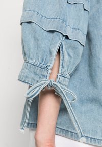 Denimskjorta i ljusblå färg med uppskjutna ärmar och justerbara snörningar med tofsar. Tyget har en mjuk, strukturerad yta.